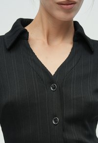 Μαύρο πουκάμισο με κουμπιά, φτιαγμένο από υφή με ριγέ ύφασμα. Διαθέτει δομημένο γιακά, ντεκολτέ σε σχήμα V και λεπτομέρεια με ένα μόνο κουμπί.