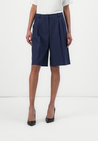 Navy pleated shorts gjorda av lättviktsmaterial, med hög midja, bred ben-design och pressade veck. Bärs med svarta högklackade skor.