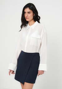 Λευκό πουκάμισο με μακριά μανίκια, γιακά και μπροστινή τσέπη, σε συνδυασμό με μια φούστα σε σκούρο μπλε χρώμα με ελαφρύ σχεδιασμό τύπου wrap. Λείο ύφασμα.