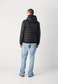 Sort pufferjakke med hætte, ribbede manchetter og quiltet tekstur. Kombination med lyseblå jeans og brune sko.