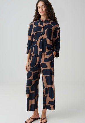 Donna in piedi che indossa una camicia morbida marrone e nera con motivo astratto e pantaloni larghi abbinati con infradito marroni.