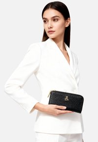 Portafoglio in pelle nera con finitura testurizzata, cerniera dorata e dettaglio con logo. Tenuto in mano su uno sfondo di blazer bianco.