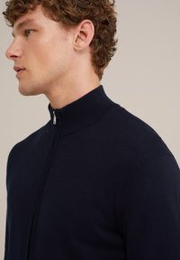 Mörkblå zip-up tröja, ribbad hög krage, figurnära design, slät textur, detaljerad sömnad i sidorna, minimal reflekterande dragkedja.