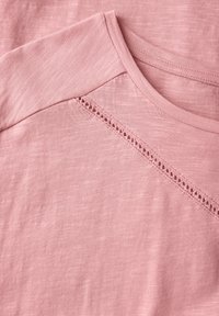 Světle růžové tričko, měkká tkanina s jemnou strukturou, má zaoblený výstřih a krajkové detaily na ramenním švu.