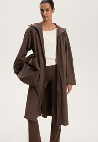 Caban lung cu glugă de culoare maro, realizat dintr-un material neted, având un fermoar în față și două deschideri laterale. O geantă maro însoțitoare este ținută sub braț.