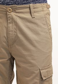 Pantaloni cargo beige con una finitura testurizzata e stropicciata, dotati di chiusura con bottone, passanti per la cintura e tasche laterali.