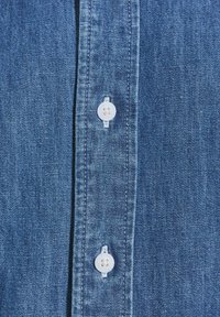 Camicia in denim con placca di bottoni, due bottoni bianchi e tessuto blu testurizzato. Design semplice, senza modelli o accenti visibili.