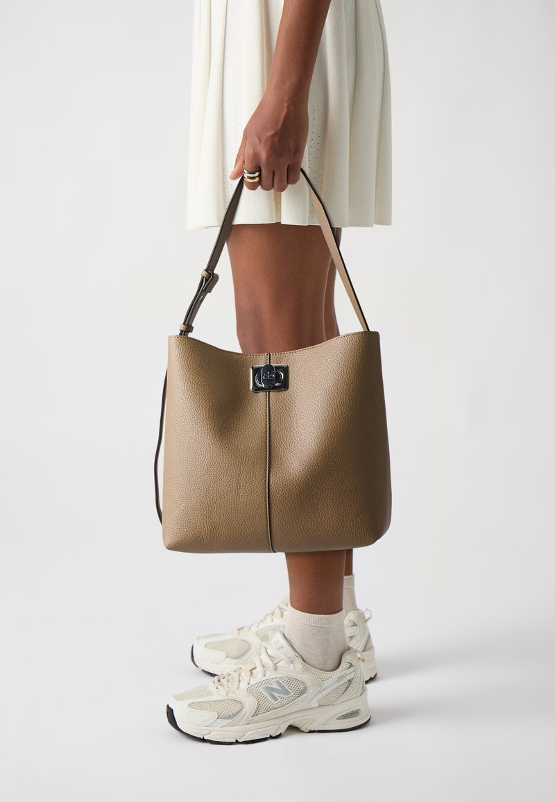 Personne portant des baskets blanches et une jupe blanche tient un grand sac à main en cuir marron par sa bandoulière sur un fond uni.