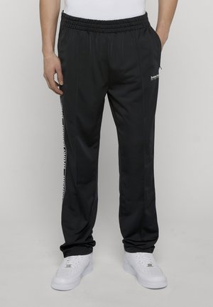 Sorte træningsbukser med elastisk taljebånd, hvidt logo på siden, båret med hvide sneakers og hvid top, hænder afslappet ved siden.