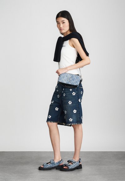 Denim shorts met witte bloemenborduursel, gecombineerd met een wit tanktop en een zwarte trui over de schouders. Blauwe tas met patroon en denim sandalen.