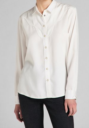 Witte blouse met lange mouwen met drukknopen en een subtiel gebogen inzetstuk, gedragen met zwarte broek.