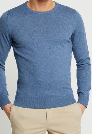 Homme portant un pull bleu ajusté à manches longues avec un col rond, des poignets et un ourlet côtelés, associé à un pantalon beige, les mains dans les poches.