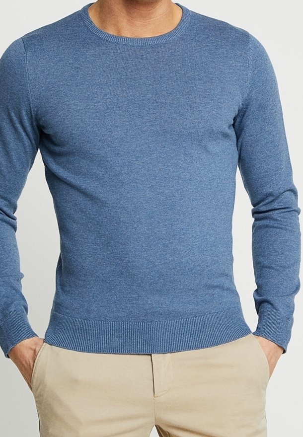 Homme portant un pull bleu ajusté à manches longues avec un col rond, des poignets et un ourlet côtelés, associé à un pantalon beige, les mains dans les poches.