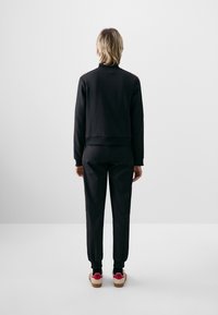 Črna majica s kapuco in usklajene hlače, s skrajšanim dizajnom. Čevlji imajo belo podlago z rdečimi detajli in teksturirano podplat.