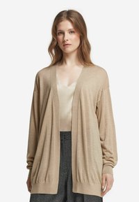 Beige Strickjacke aus leichtem Material, mit V-Ausschnitt, offenem Front und gerippten Bündchen. Kombiniert über einem hellen Oberteil und gemusterten Hosen.