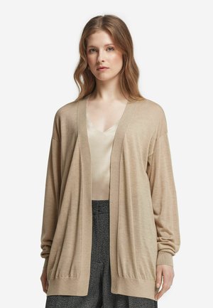 Cardigan beige en maille légère, avec un design en V, une ouverture frontale et des poignets côtelés. Porté par-dessus un haut clair et un pantalon à motifs.