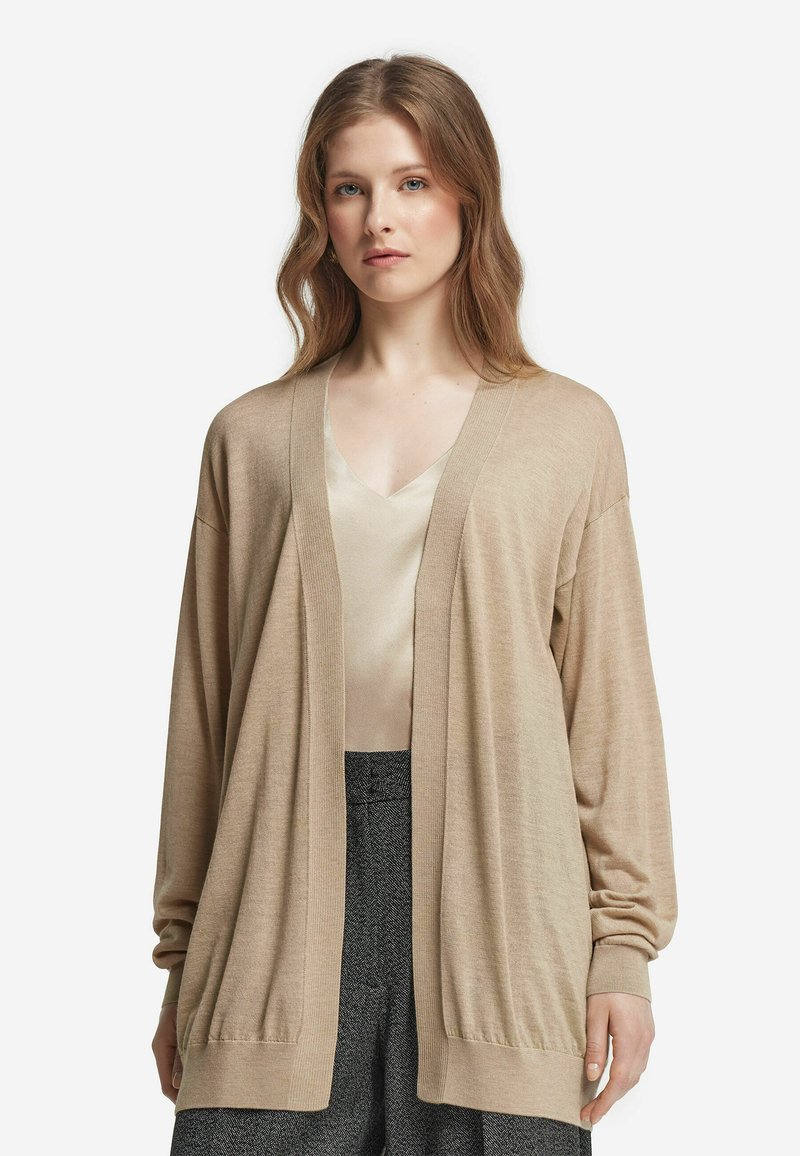 Beige Strickjacke aus leichtem Material, mit V-Ausschnitt, offenem Front und gerippten Bündchen. Kombiniert über einem hellen Oberteil und gemusterten Hosen.