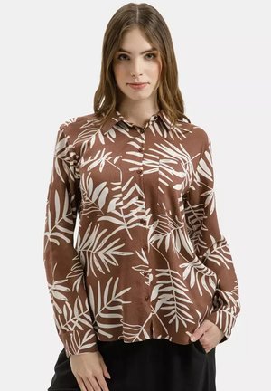 Kobieta ubrana w brązową koszulę z guzikami, ozdobioną białym tropikalnym wzorem liści, stojąca z jedną ręką w kieszeni na gładkim tle.