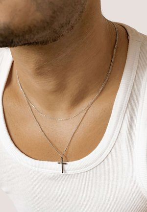 Silberne Kettenhalsketten übereinander getragen, wobei eine eine schwarze Kreuzanhänger hat. Getragen über einem weißen gerippten Tanktop. Glatte Hauttextur sichtbar.