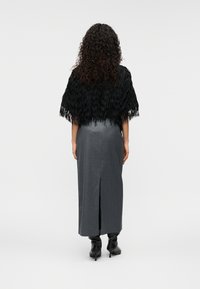 Cape noire à franges portée sur une jupe grise fendue sur le côté. Le mannequin porte des bottes à talons noires. Tissu lisse avec une silhouette structurée.