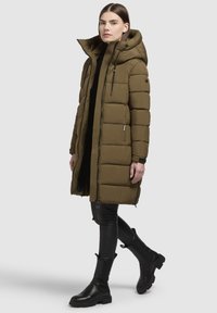 Abrigo acolchado en verde oliva con capucha, textura de acolchado, cierre delantero y bolsillos laterales. Lleva puesto un top negro y pantalones negros ajustados.