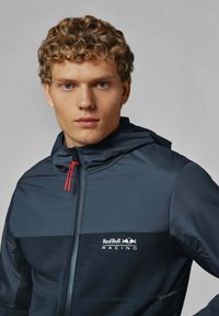 Muška jakna s kapuljačom od tamnoplave tkanine. Ima prednji zip, dva bočna džepa i kontrastnu crvenu vezicu. Prikazan Red Bull Racing logo.
