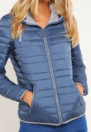 Femme portant une veste matelassée bleue zippée avec un bord gris et une capuche, les mains dans les poches latérales zippées, associée à un jean bleu.