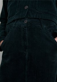 Ensemble veste et jupe en velours côtelé vert foncé avec nervures verticales, deux boutons et poches latérales. Le tissu texturé ajoute de la profondeur.