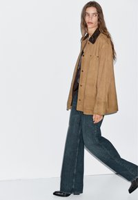 Giacca in suede marrone con colletto scuro, abbinata a jeans in denim a gamba larga. Il modello indossa stivaletti neri alla caviglia, in piedi su uno sfondo bianco.