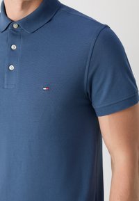 Homme portant un polo bleu à manches courtes avec trois boutons blancs et un petit logo rectangulaire rouge, blanc et bleu sur la poitrine.