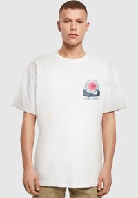 Camiseta de algodón blanca con un diseño gráfico de un sol, olas y el texto "SUNSET CHARM" en el lado izquierdo del pecho. Mangas cortas y anchas.