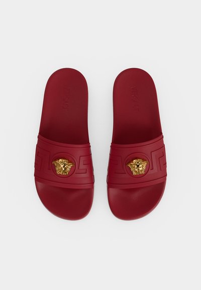 Paire de sandales slides rouges avec motifs embossés et décorations tête de Méduse dorées sur les brides, vues de dessus sur fond blanc.