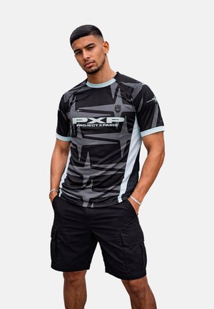 Hombre con camiseta gráfica de manga corta en negro y gris con el logo "PXP Project X Paris" y pantalones cortos cargo negros, posando con las manos en los bolsillos.