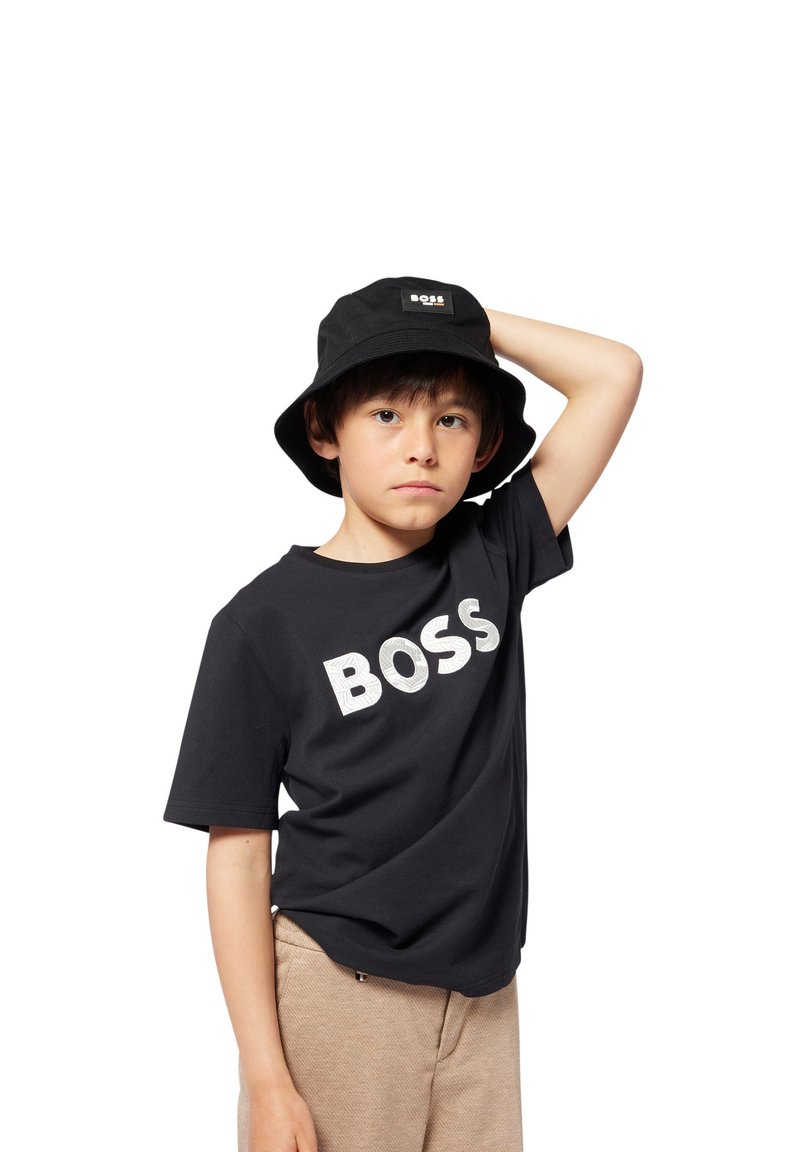 Cappello a secchiello nero con patch logo, abbinato a una maglietta nera a maniche corte con la parola "BOSS" in lettere bianche in grassetto e strutturate, e pantaloni corti beige.