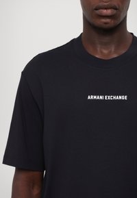 Homme portant un t-shirt noir Armani Exchange avec un logo blanc de la marque sur la poitrine gauche, visible du cou jusqu'au milieu du torse.