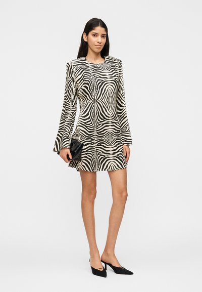 Γυναίκα που φοράει μαύρο και άσπρο μίνι φόρεμα με μακριά μανίκια και σχέδιο ζέβρας, μαύρα μυτερά τακούνια και κρατάει μια μικρή μαύρη τσαντούλα χειρός.