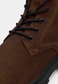 Stivale in pelle marrone con finitura texture, lacci neri e occhielli metallici. Presenta un colletto imbottito e una suola in gomma robusta.