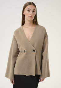 Cardigan avvolgente in maglia beige con scollo a V profondo, maniche a campana oversize e chiusura a doppio bottone nero. Texture liscia, vestibilità comoda.