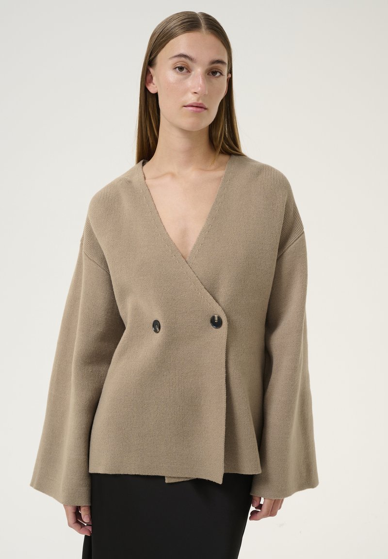 Cardigan avvolgente in maglia beige con scollo a V profondo, maniche a campana oversize e chiusura a doppio bottone nero. Texture liscia, vestibilità comoda.