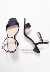 Námořnické saténové sandály na tenkých páscích se štíhlým designem, úzkým páskem kolem kotníku a dekorativním krystalovým detailem na patě. Styl s otevřenou špičkou.