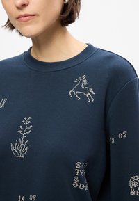 Σκούφος ναυτικού μπλ ε με στρογγυλή λαιμόκοψη, διαθέτει ασημένια κεντήματα με άλογο, φυτικά μοτίβα και αριθμούς. Ύφασμα από κράμα βαμβακιού με λεία υφή.