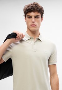 Polo beige avec col, arborant un petit logo sur la poitrine et des finitions contrastées sur les manches. Le modèle tient une veste noire.