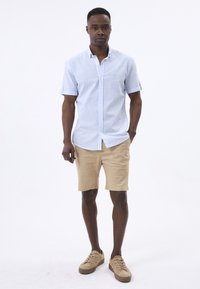 Camisa de manga corta a rayas azul claro y blanca, pantalones cortos beige y zapatillas beige. Diseño sencillo con collar abotonado y mangas arremangadas.