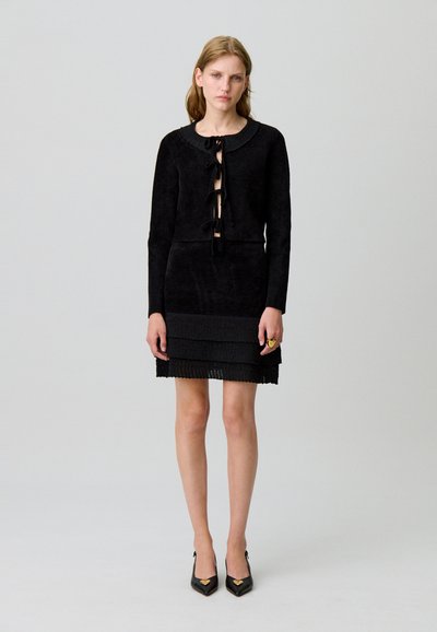 Vestido negro texturizado con una chaqueta corta que presenta un escote redondo y lazos en la parte frontal. El vestido incluye acentos de tela en capas en el dobladillo.