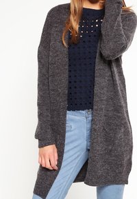 Mörkgrå cardigan med öppen framsida, långa ärmar och en mjuk textur, bärs över en marinblå topp med ett perforerat mönster.