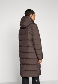 Brun pufferjakke med hette, kjennetegnet av quiltede, polstrede seksjoner. Lang design som når under knærne. Glatt stofftekstur.