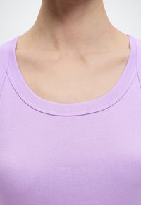 Λαιμός και άνω μέρος του θώρακα ενός ατόμου που φοράει ανοιχτό μωβ ριγωτό αμάνικο τοπ με στρογγυλή λαιμόκοψη σε λευκό φόντο.