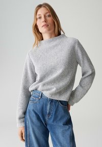 Donna bionda che indossa un maglione grigio a maniche lunghe con trama e jeans blu larghi, in piedi contro uno sfondo chiaro uniforme.