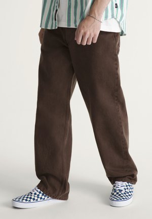 Person trägt braune lockere Hose, blau-weiß karierte Sneakers und ein grün gestreiftes Hemd mit einem Armband am linken Handgelenk.