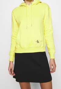 Osoba w żółtej bluzie z kapturem, posiadającej kieszeń z przodu oraz czarnym logo "cK", zestawiona z czarną spódnicą, stojąca na tle jednolitym.